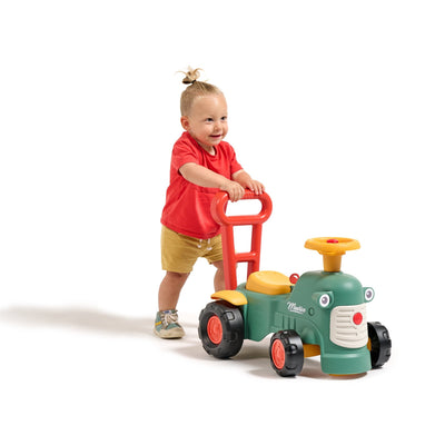 A toddler in a red shirt, learning to walk, pushes the TP Toys Falk Maurice Vintage Baby Green Tractor—featuring large wheels and a steering wheel.