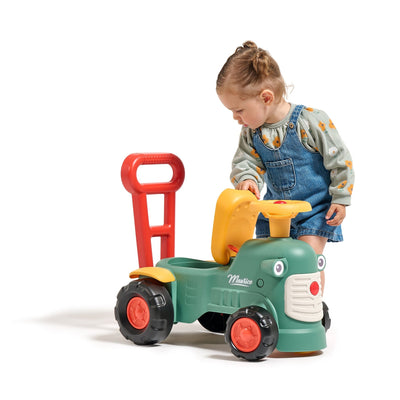 A young child in a gray shirt and denim overalls stands next to the TP Toys Falk Maurice Vintage Baby Green Tractor, lifting its seat.