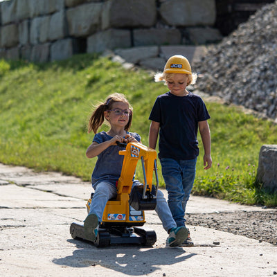 A girl rides the TP Toys Falk JCB Fully Articulated Digger with Opening Seat and Helmet, while a boy in a yellow hard hat walks beside her on a paved path, both enjoying construction role play outdoors.