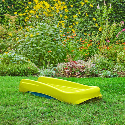 The TP Toys PalPlay Green & Blue Plastic Folding First Slide is placed on green grass in a garden, with flowering plants and dense greenery in the background.
