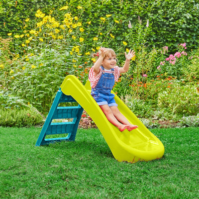 A young child in blue overalls and a striped shirt slides down the TP Toys PalPlay Green & Blue Plastic Folding First Slide in a garden with green grass and flowering plants.