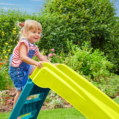 A young child in denim overalls and a striped shirt climbs the steps of the TP Toys PalPlay Green & Blue Plastic Folding First Slide in a garden.