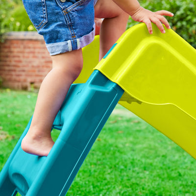 A young child in denim shorts climbs the TP Toys PalPlay Green & Blue Plastic Folding First Slide outdoors, with green grass and a brick wall in the background.