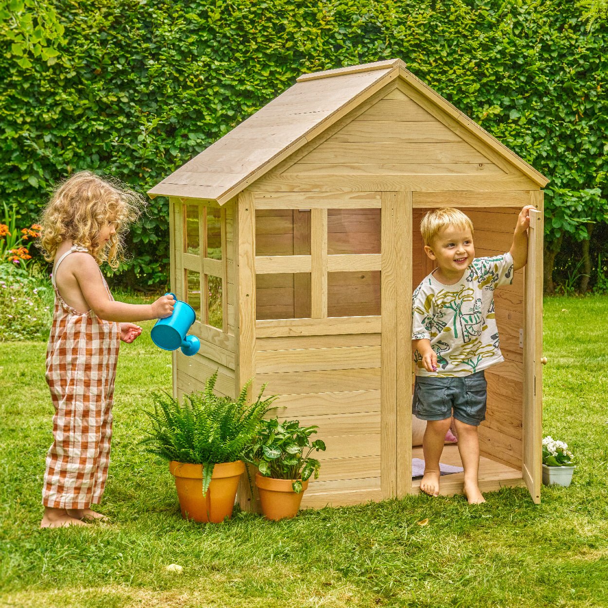 Two children play outdoors with the Buttercup Cottage Wooden Playhouse with Floor by TP Toys (FSC® certified); one waters potted plants as the other stands in the doorway.