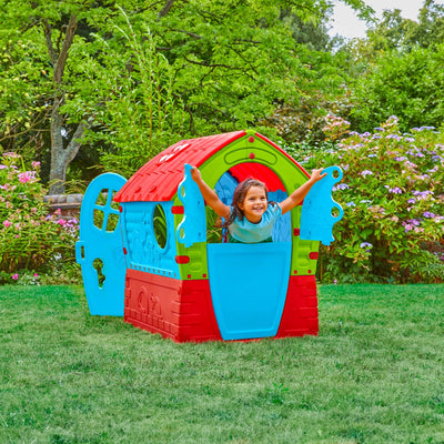 A young child smiles while playing inside the TP Toys Palplay Plastic Dream Playhouse, a colorful UV-resistant playhouse with open doors, set on grass and surrounded by trees and flowers.