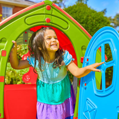 A smiling young girl stands in the doorway of the TP Toys Palplay Plastic Dream Playhouse, a colorful UV-resistant outdoor playhouse, on a sunny day.