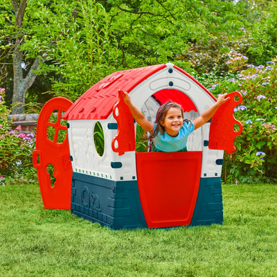 A young child smiles while leaning out of the window of a TP Toys Palplay Plastic Dream Playhouse, featuring an open door and UV-resistant colors, set on green grass in a garden.