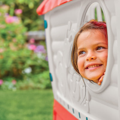 A young child smiles and looks out of a window in the TP Toys Palplay Plastic Dream Playhouse, featuring UV-resistant plastic, with a blurred green garden behind—ideal for indoor or outdoor play.