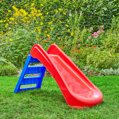 The TP Toys PalPlay Red & Blue Plastic Folding First Slide, featuring blue steps, stands on green grass among flowers and plants. This compact slide is ideal for indoor or outdoor play.