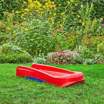 The TP Toys PalPlay Red & Blue Plastic Folding First Slide lies flat on the green grass in a garden with flowers and plants in the background.