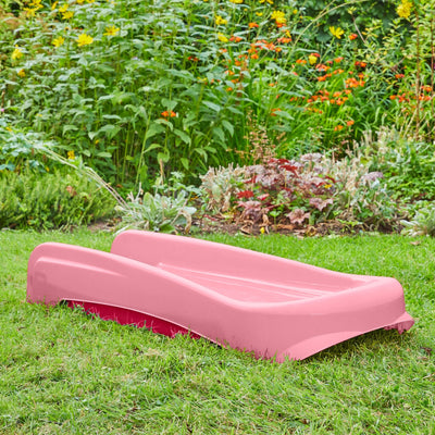 The TP Toys PalPlay Pink & White Plastic Folding First Slide is set on garden grass, surrounded by green plants and colorful flowers in the background.
