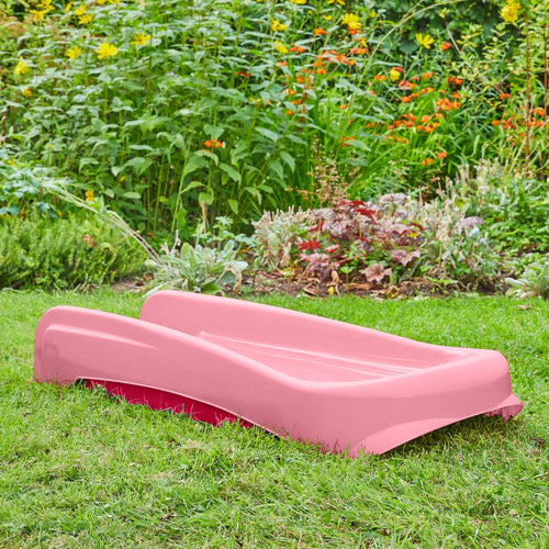 The TP Toys PalPlay Pink & White Plastic Folding First Slide is set on garden grass, surrounded by green plants and colorful flowers in the background.