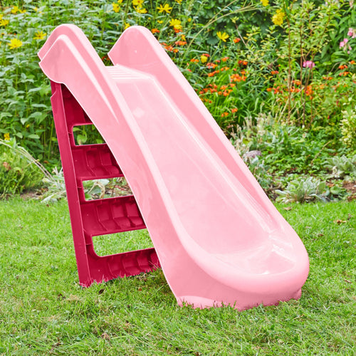 The TP Toys PalPlay Pink & White Plastic Folding First Slide is set up on green grass in a garden, with its bright colors contrasting the surrounding colorful flowers.