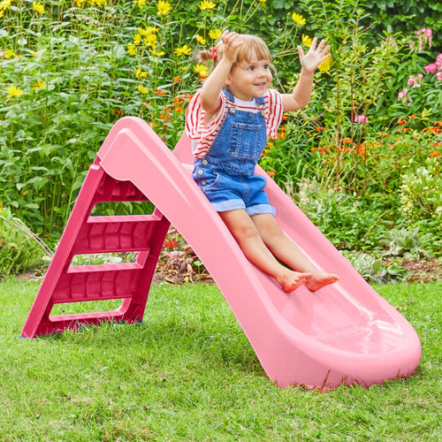 A young child in denim overalls enjoys the TP Toys PalPlay Pink & White Plastic Folding First Slide on green grass in a garden with flowering plants.