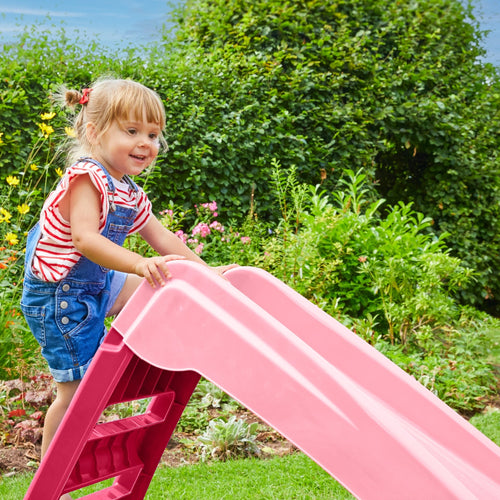 A young child in denim overalls and a striped shirt climbs the TP Toys PalPlay Pink & White Plastic Folding First Slide in a garden with green bushes and flowers.