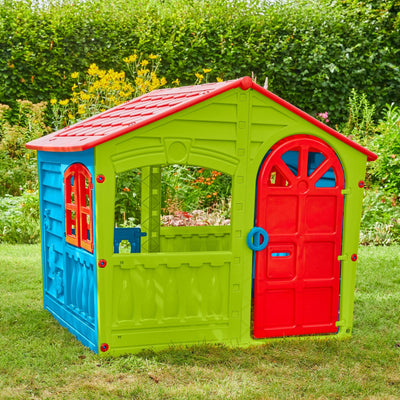 The TP Toys Palplay Plastic Playhouse of Fun features a bright red door, blue window panels, and green walls—perfect for indoor or outdoor fun, set on garden grass amidst flowers and hedges.