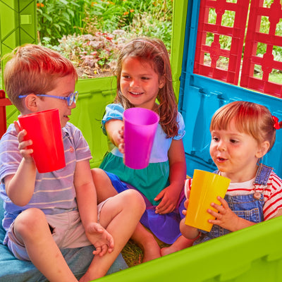 Three young children sit inside the TP Toys Palplay Plastic Playhouse of Fun, each holding red, purple, and yellow plastic cups, smiling at each other.