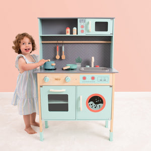 A young child stands next to the TP Toys Owl & Fox Wooden Imagination Kitchen & Baking Set, smiling while pretending to cook with its oven, sink, microwave, and toy accessories for creative pretend play.