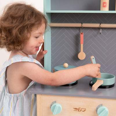 A young child smiles while playing with the TP Toys Owl & Fox Wooden Imagination Kitchen Set—FSC® certified—stirring a pot with a wooden spoon.