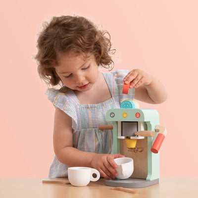 A young child in a pastel dress enjoys pretend play with the TP Toys Owl & Fox Wooden Coffee Machine – FSC® certified, placing a pretend pod inside and holding a cup under the spout.