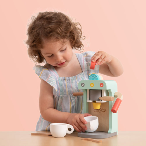 A young child in a pastel dress enjoys pretend play with the TP Toys Owl & Fox Wooden Coffee Machine – FSC® certified, placing a pretend pod inside and holding a cup under the spout.