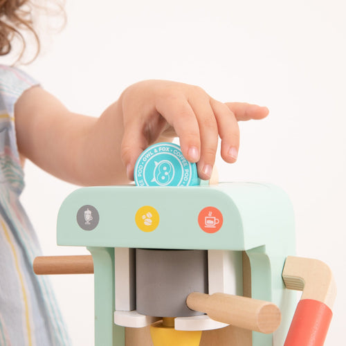 A child inserts a blue wooden coffee pod into the TP Toys Owl & Fox Wooden Coffee Machine—FSC® certified—adorned with colorful icons, inspiring imaginative pretend play.