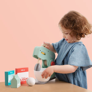 A young child in a blue dress enjoys pretend play at a wooden table with the TP Toys Owl & Fox Wooden Baking Set - FSC® certified, featuring a toy mixer, bowl, and boxes labeled flour, sugar, and milk.