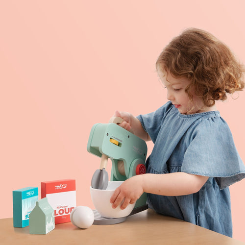 A young child in a blue dress enjoys pretend play at a wooden table with the TP Toys Owl & Fox Wooden Baking Set - FSC® certified, featuring a toy mixer, bowl, and boxes labeled flour, sugar, and milk.
