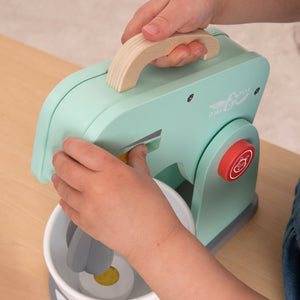 A child plays with the mint green TP Toys Owl & Fox Wooden Baking Set, holding the mixer handle while blending toy food in a white bowl on a wooden play kitchen set.
