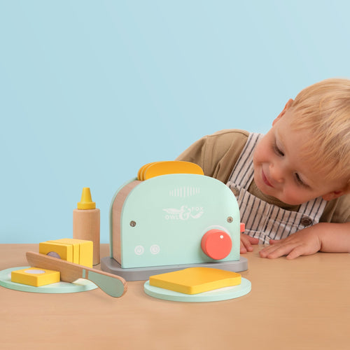 A young child admires the TP Toys Owl & Fox Wooden Toaster Set—an FSC® certified pastel playset with toy bread, butter, knife, and bottle—perfect for pretend play and Montessori activities.