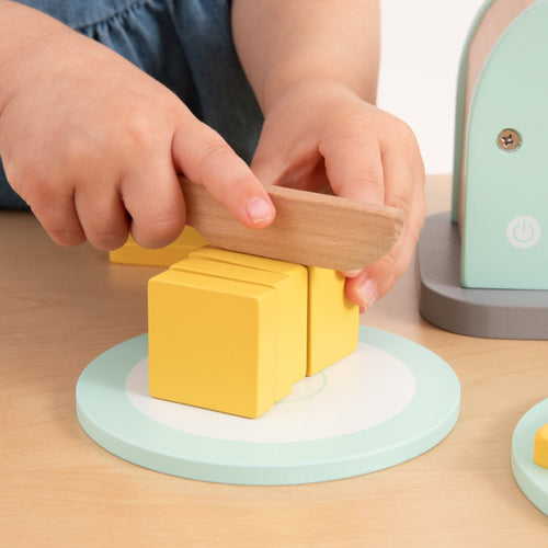 A child enjoys pretend play with the Owl & Fox Wooden Toaster Set from TP Toys, using FSC® certified wooden pieces on a pastel-colored cutting board for charming Montessori-inspired fun.