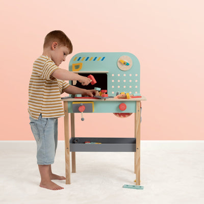 A young boy plays with the TP Toys Owl & Fox Wooden Workbench Set – FSC® certified, pretending to build and use the included tools and accessories in a brightly lit room with a light pink wall.