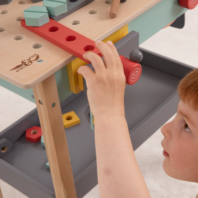 A child plays with the TP Toys Owl & Fox Wooden Workbench Set (FSC® certified), placing a red holed piece onto the bench among geometric blocks and pretend play accessories.