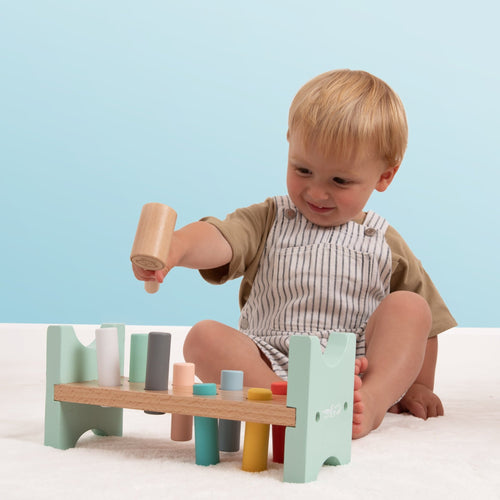 A young child sits on the floor using the TP Toys Owl & Fox Wooden Hammer Bench—FSC® certified—to build hand-eye coordination, playing with its pastel colors against a light blue background.