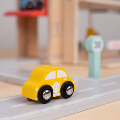 A small yellow toy car is on a gray toy road near a green “31” sign, with the TP Toys Owl & Fox Wooden Multi-storey Garage with Vehicle Transporter (FSC® certified) blurred in the background—ideal for pretend play or as a Montessori toy accessory.