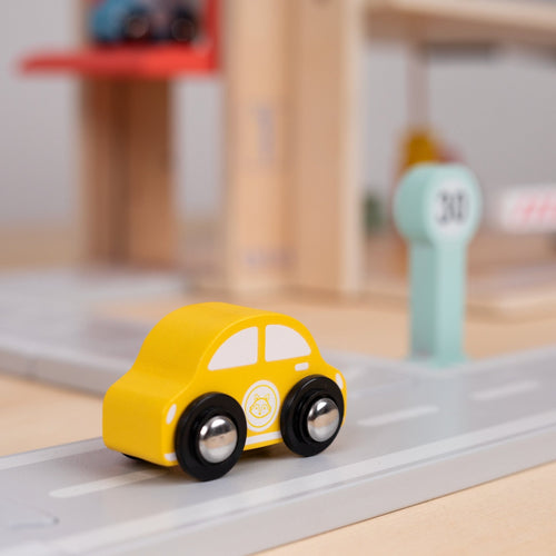 A small yellow toy car is on a gray toy road near a green “31” sign, with the TP Toys Owl & Fox Wooden Multi-storey Garage with Vehicle Transporter (FSC® certified) blurred in the background—ideal for pretend play or as a Montessori toy accessory.
