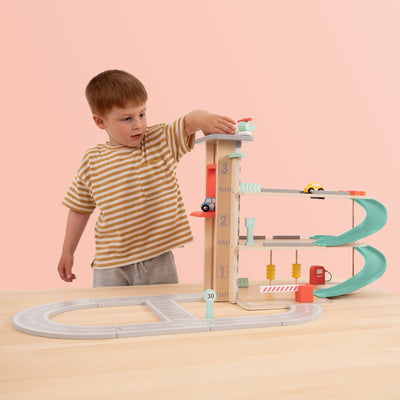 A young boy in a striped shirt plays with the TP Toys Owl & Fox Wooden Multi-storey Garage with Vehicle Transporter (FSC® certified) on a light-colored table, enjoying creative pretend play against a peach background.