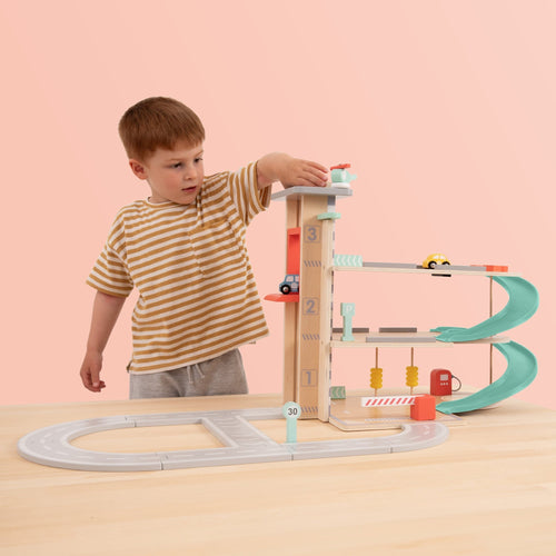 A young boy in a striped shirt plays with the TP Toys Owl & Fox Wooden Multi-storey Garage with Vehicle Transporter (FSC® certified) on a light-colored table, enjoying creative pretend play against a peach background.
