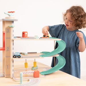 A young child plays with the TP Toys Owl & Fox Wooden Multi-storey Garage with Vehicle Transporter (FSC® certified), driving cars on ramps and levels, as a toy helicopter and other accessories spark imaginative fun.