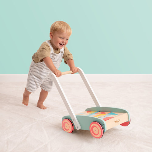 A toddler pushes the TP Toys Owl & Fox Wooden Baby Walker and Blocks (FSC® certified) across a light carpet in front of a mint green wall.