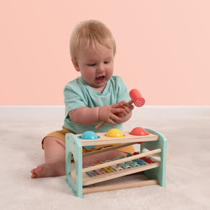 A baby sits on the floor, smiling as they play with the TP Toys Owl & Fox Wooden Xylophone Bench—an FSC® certified Montessori toy—using a mallet to build fine motor skills.