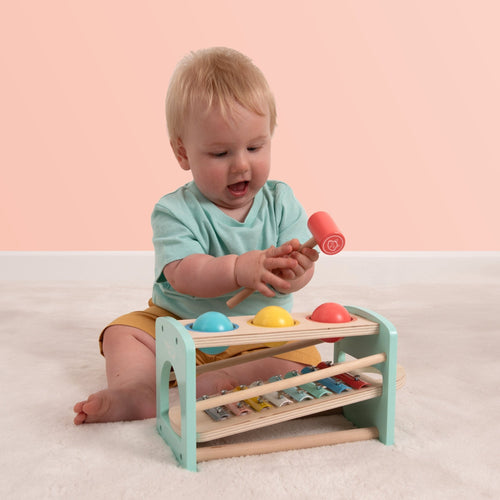 A baby sits on the floor, smiling as they play with the TP Toys Owl & Fox Wooden Xylophone Bench—an FSC® certified Montessori toy—using a mallet to build fine motor skills.