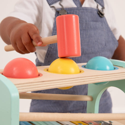 A child in blue overalls uses a mallet to strike a yellow ball on the Owl & Fox Wooden Xylophone Bench by TP Toys, an FSC® certified Montessori toy that promotes fine motor skills with colorful, engaging wooden elements.