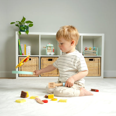 A young child sits on the floor playing imaginatively with TP Toys Owl & Fox Wooden Pancake Set - FSC® certified kitchen utensils and toy food, with shelves and baskets in the background.