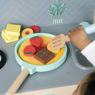 A child enjoys imaginative play with the TP Toys Owl & Fox Wooden Pancake Set - FSC® certified, cooking chocolate, strawberries, and butter in their play kitchen.