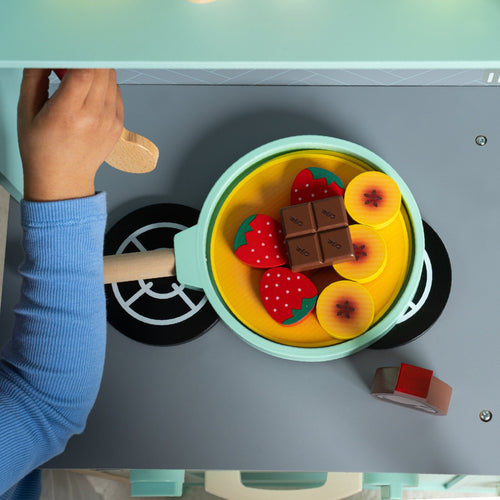 A child’s hand holds a wooden spoon beside the TP Toys Owl & Fox Wooden Pancake Set—FSC® certified—with play food shaped like strawberries, chocolate, and fruit, perfect for imaginative play.