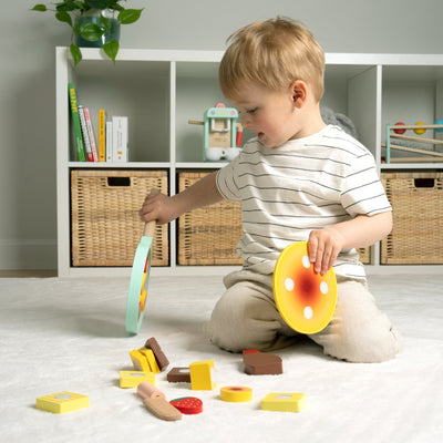 A young child enjoys imaginative play with the TP Toys Owl & Fox Wooden Pancake Set (FSC® certified) in a bright, organized room, sitting on a carpet with shelves and baskets in the background.