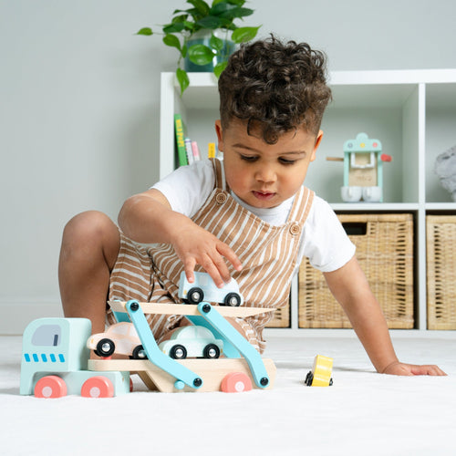 A young child in striped overalls plays on the floor with the TP Toys Owl & Fox Wooden Multi-storey Garage with Vehicle Transporter (FSC® certified), surrounded by bright light and pretend play accessories in an organized room.