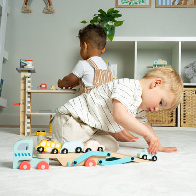 Two young children play on the floor with TP Toys’ Owl & Fox Wooden Multi-storey Garage with Vehicle Transporter—FSC® certified—in a bright, organized playroom, encouraging imaginative pretend play amid shelves and decor.