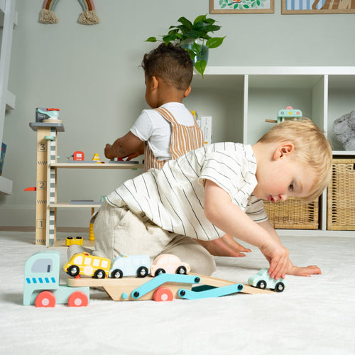 Two young children play on the floor with TP Toys’ Owl & Fox Wooden Multi-storey Garage with Vehicle Transporter—FSC® certified—in a bright, organized playroom, encouraging imaginative pretend play amid shelves and decor.